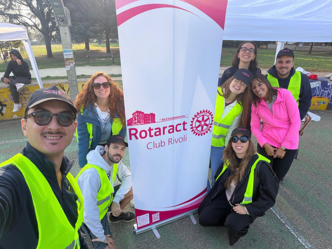 Il gruppo del Rotaract Rivoli con i gilet gialli in un selfie davanti al roll-up del club, in un parco.